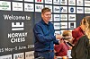 Mann deler ut sjokolade ved en Norway Chess-stand foran en vegg med sponsorlogoer.