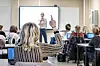 Lærer foran en skjerm i et klasserom mens elever sitter ved pultene, og en elev rekker opp hånden.