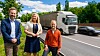 Tre personer står ved en vei, med en lastebil i bevegelse bak dem.