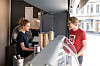 To personer snakker ved disken i en kafé med kaffekopper og vindu ut mot gaten.