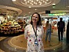 Woman standing in an airport concourse near a Vitasapotek pharmacy with shoppers and store displays behind her.