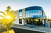 Moderne kontorbygg i glass med blå logo, trær og sollys ved inngangspartiet.