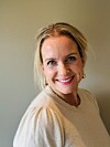 Smilende kvinne med lyst hår og lys genser fotografert innendørs mot en beige vegg.