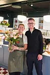 Carmen og Frank foran en cafédisk. Hun har forkle og holder i en vase med blomster. Begge to smiler og er blide.