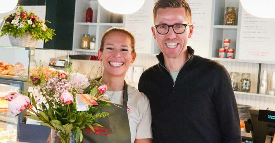 Carmen og Frank foran en cafédisk. Hun har forkle og holder i en vase med blomster. Begge to smiler og er blide.